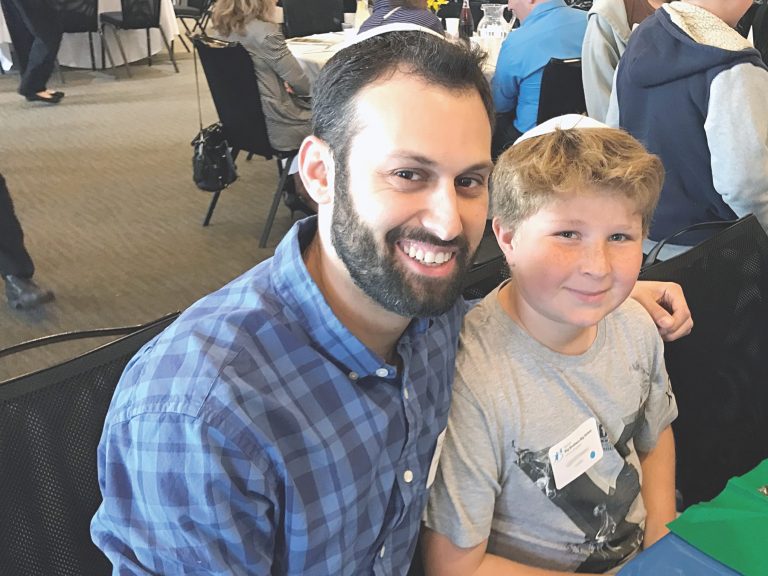 Two people, an adult and a child, sit together at an event. The adult wears a blue plaid shirt and the child a gray T-shirt. Both wear kippahs and smile at the camera, embodying the spirit of a Jewish mentorship program.