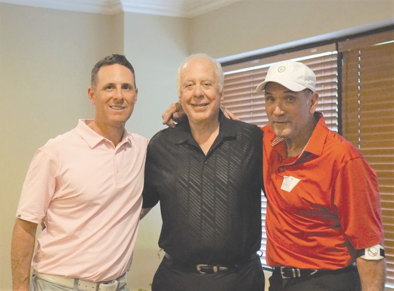 Three men standing shoulder to shoulder indoors, smiling at the camera. They are wearing casual polo shirts and one is wearing a white baseball cap, likely celebrating their involvement in a Jewish mentorship program.