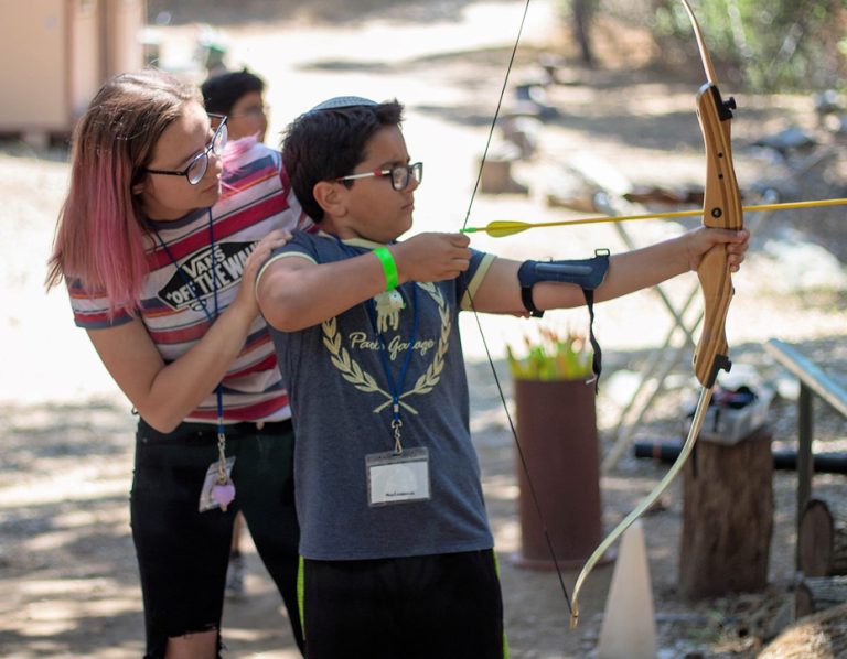 A woman assists a child with pink-tinted hair as he aims a bow and arrow outdoors at a Jewish summer camp. Both are wearing glasses and name tags, and the child is equipped with an arm guard and wristband.