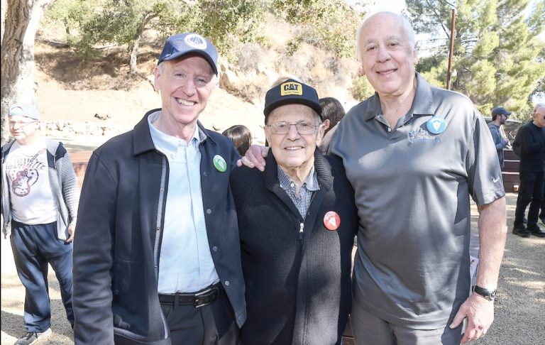 Three older men are standing together outdoors, smiling at the camera. Two of them are wearing jackets, and the man in the middle is wearing a black cap. Trees and people can be seen in the background, creating a warm atmosphere reminiscent of a Jewish summer camp.