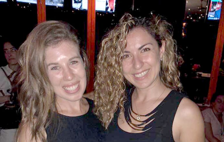 Two women with long, wavy hair smile at a dimly lit indoor event with a few people and television screens in the background. Both are dressed in black tops, possibly enjoying a gathering related to a Jewish mentorship program.