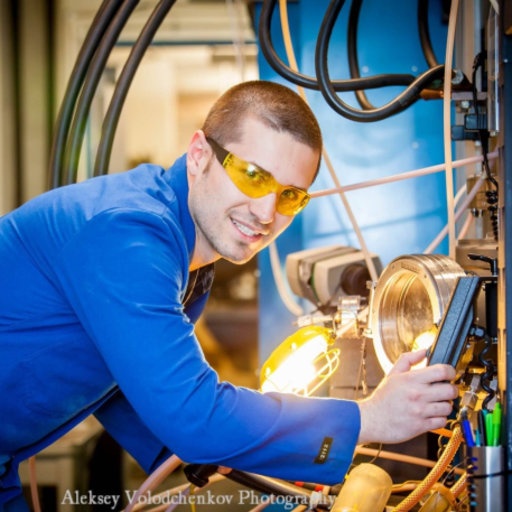 A man in a blue lab coat and yellow safety glasses, reminiscent of a mentor from a Jewish youth program, operates machinery in an industrial setting, adjusting controls with his right hand.