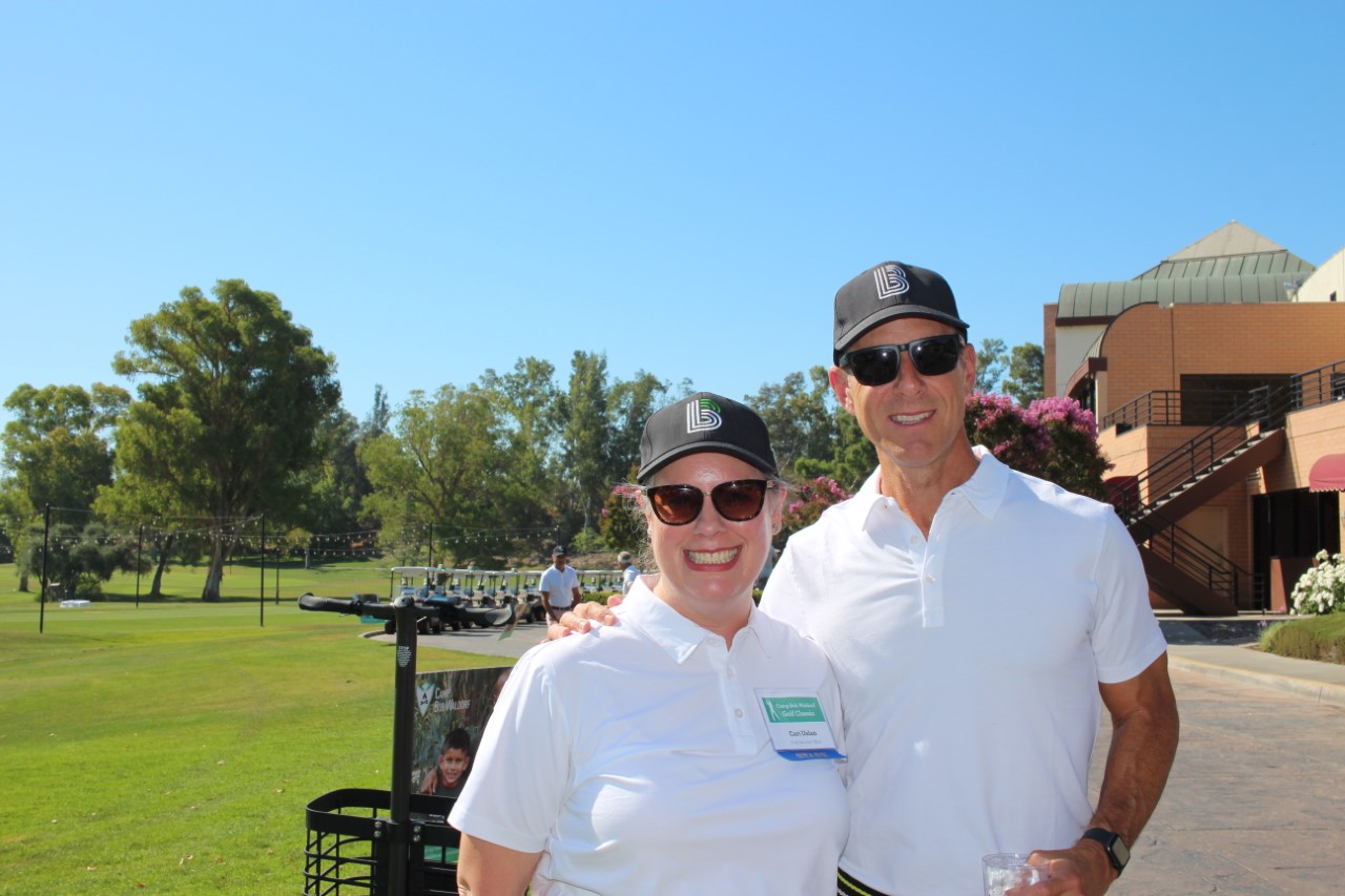 Two people wearing white polo shirts and baseball caps smiling at a golf course. One person is wearing a name tag, and trees and buildings are visible in the background on a sunny day, likely enjoying a break from their Jewish mentorship program.