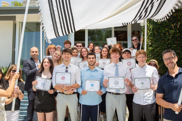 A group of young people and adults pose outdoors, several holding certificates, under a canopy in front of a modern building.