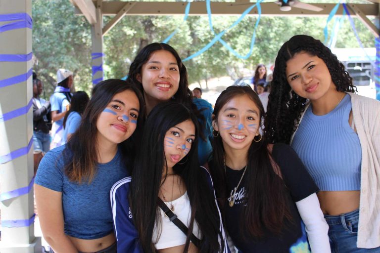 Five young women with blue face paint and casual clothing smile at the camera under a gazebo decorated with blue streamers. Trees are visible in the background.