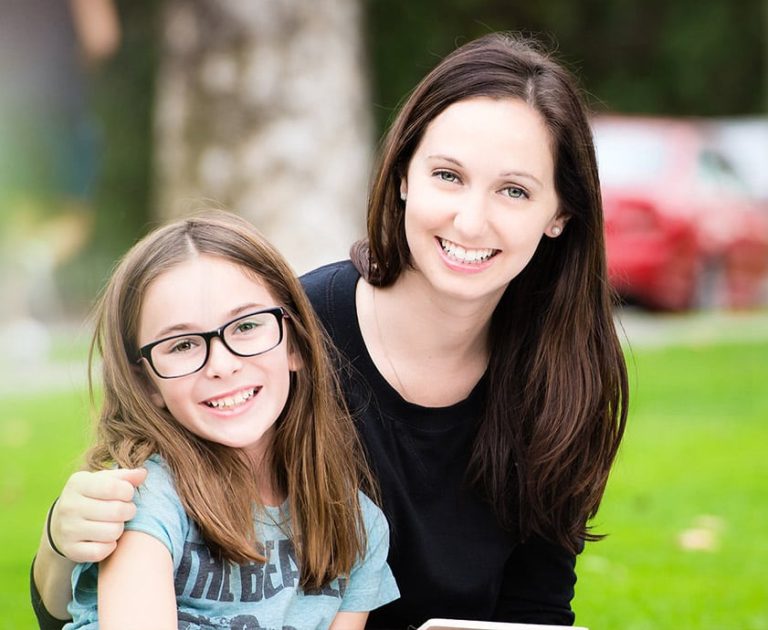 A woman and a young girl with long brown hair and glasses sit closely together on the grass, both smiling at the camera.